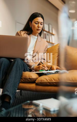 Donna d'affari concentrata che scrive in diario mentre si siede con un computer portatile sul divano in ufficio Foto Stock
