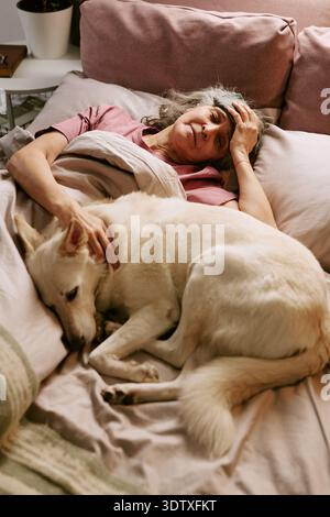 Donna caucasica anziana sdraiata nel letto cane da compagnia e sorridente al mattino Foto Stock
