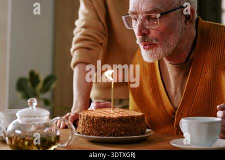 Senior Caucasico Man Blowing Out Candle on Birthday Cake festeggia al chiuso Foto Stock