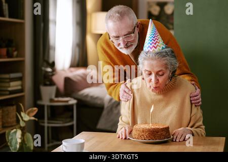 Donna caucasica anziana che soffia la candela per la torta di compleanno con l'uomo anziano che guarda Foto Stock