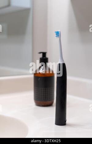 Uno spazzolino sonico nero in un bagno bianco, con un dispenser nero di sapone e crema posizionato dietro di esso. Design moderno ed elegante Foto Stock