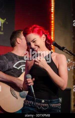 Giovane donna dai capelli rossi brillanti che ride tenendo in mano un microfono sul palco con chitarrista in sottofondo, luce calda Foto Stock