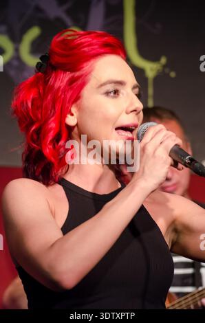 Cantante donna con capelli rossi vivaci che si esibisce sul palco, tenendo un microfono a metà canzone, faccia espressiva, sfondo scuro con testo sfocato e musicia Foto Stock