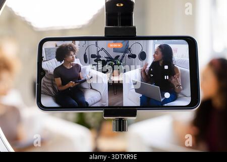 Le donne afroamericane ospitano registrazioni sullo schermo del telefono in soggiorno con abiti casual con microfoni Foto Stock