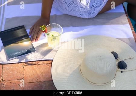 Il cappello da sole in paglia intrecciata si appoggia sul patio in mattoni a bordo piscina con occhiali da sole, asciugamano, libro, bevanda Foto Stock