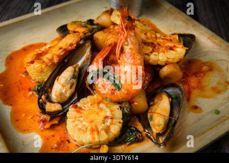 Un piatto di pesce saltato in padella piccante con gamberi, cozze e baby corn, servito con una ricca e saporita salsa su un piatto di legno rustico. Foto Stock