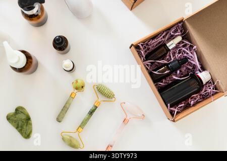 Gli strumenti per il massaggio facciale vengono disposti su un tavolo bianco con rulli di giada, gua Sha e bottiglie color ambra Foto Stock