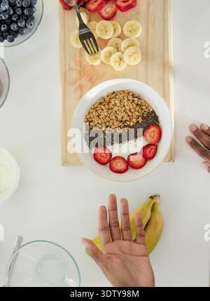 Uomo afro-americano che raggiunge e monta la ciotola di yogurt con granola e chia su un tavolo bianco Foto Stock