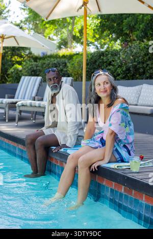 Coppia afroamericana seduta a bordo piscina immergendo i piedi in acqua limpida con sfumature e cocktail Foto Stock