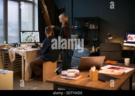 Giovane uomo adulto caucasico seduto alla scrivania che lavora al computer, mentre una donna caucasica sta accanto a lui, che punta la foto sul monitor in un moderno spazio di lavoro creativo Foto Stock