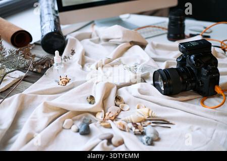 Scena STILL Life con fotocamera digitale professionale collegata tramite cavo, conchiglie sparse, ciottoli, piante secche, bottiglia contagocce e tastiera per computer su tessuto drappeggiato Foto Stock
