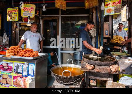 Uno stand di cibo di strada a nuova Delhi dove un venditore di patatine fritte jalebi in un grande vaso, con dolci tradizionali e poster esposti in lontananza. Foto Stock