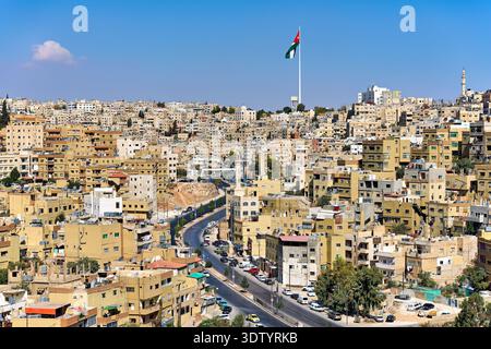 Amman mostra un'ampia vista di edifici, strade e la bandiera giordana che sventola sulla collina. La gente si muove attraverso questa trafficata area urbana alla luce del giorno. Foto Stock