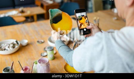 Mani di una donna che fotografa i suoi prodotti con uno smartphone in uno studio di ceramica Foto Stock