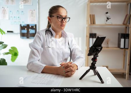 medico donna che indossa occhiali da vista e camice bianco che utilizza il telefono sul treppiede, consultando la paziente online. terapeuta che fa videocall nella sua clinica medica Foto Stock