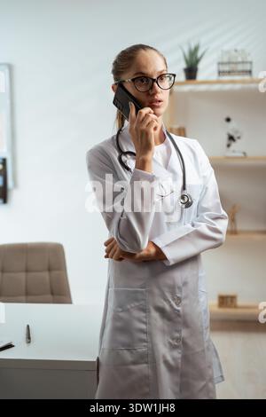 Medico di medicina generale che riceve una telefonata utilizzando lo smartphone che lavora nel suo ufficio medico, supporta i pazienti da remoto Foto Stock