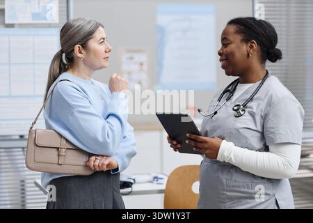 Donna caucasica di mezza età impegnata in conversazione con infermiera donna nera Foto Stock