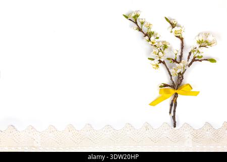 Un ramo con piccoli fiori bianchi e un nastro giallo è posizionato su una superficie bianca accanto a una finitura in pizzo, creando una configurazione semplice. Foto Stock