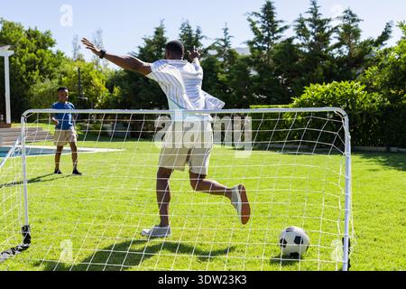 Afroamericani adulti e adolescenti che giocano a calcio in cortile, saltando la palla di blocco al pop-up goal Foto Stock