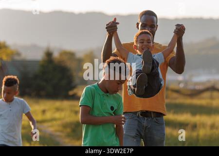 Padre e due bambini condividono risate e giocano all'aperto mentre il sole tramonta sul paesaggio creando momenti felici insieme. Foto Stock