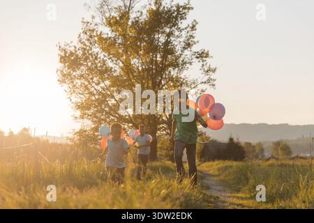 Padre e bambini corrono attraverso un campo con palloncini colorati durante il tramonto mentre si divertono insieme all'aria aperta. Foto Stock