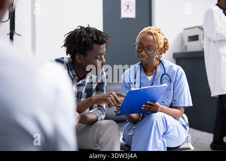 La donna medico scrive osservazioni dettagliate mentre si consulta l'uomo in un'area di accoglienza ospedaliera. Infermiera in scrub spiega informazioni sulla salute e rassicura il paziente con una discussione mirata sul trattamento. Foto Stock