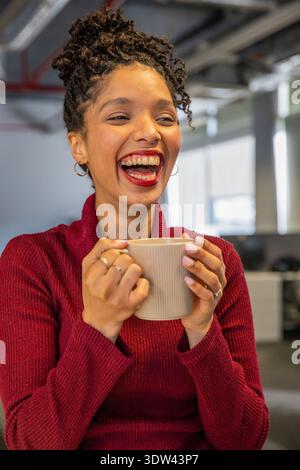 Donna afroamericana metà anni '20 che ride e tiene in mano una tazza beige in un ufficio moderno con un maglione rosso Foto Stock