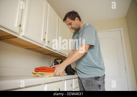 Uomo di mezza età che impilava gli asciugamani piegati nella lavanderia domestica organizzando la lavanderia pulita dopo il lavaggio durante la routine quotidiana della casa Foto Stock
