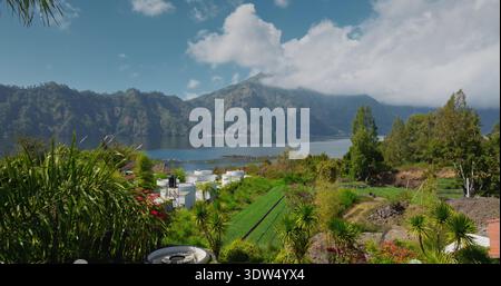 Paesaggio naturale tropicale del lago Batur con il vulcano del monte Abang parzialmente coperto da nuvole, verdi colline, terrazze agricole campi e palme sotto un cielo azzurro. Riprese di volo con drone aereo Foto Stock
