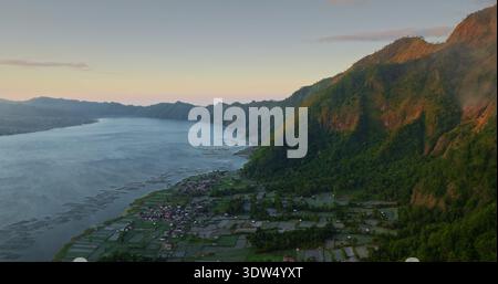 Tranquillo lago Batur con zattere d'acquacoltura, piccolo villaggio rurale e ampie risaie annidate alla base delle lussureggianti colline verdi del Monte Abang a Bali durante la tenue alba. Volo con droni per vista aerea Foto Stock