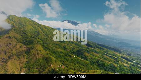 Indonesia, Bali: Lussureggianti pendii verdi del Monte Abang con basse nuvole e il lontano Monte Batur che si innalza sotto un cielo blu brillante, un suggestivo paesaggio vulcanico tropicale. Volo aereo con drone per ridurre il panorama Foto Stock