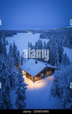 La vista aerea di un'accogliente cabina si illumina calorosamente tra pini innevati, affacciandosi sulla distesa ghiacciata di Ruka, Pohjois-Pohjanmaa, Finlandia. Foto Stock