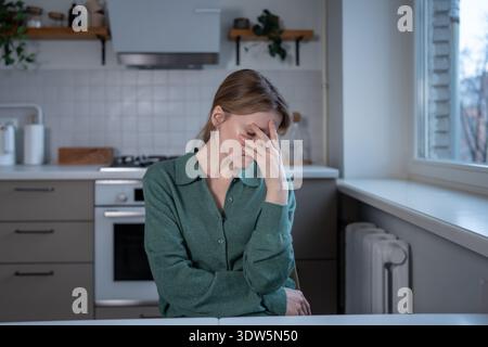 La donna stanca seduto al tavolo della cucina sente fatica a bassa energia coprire il viso con la mano. Donna assonnata stanca che tocca la testa con tensione. Esausto dallo stress Foto Stock