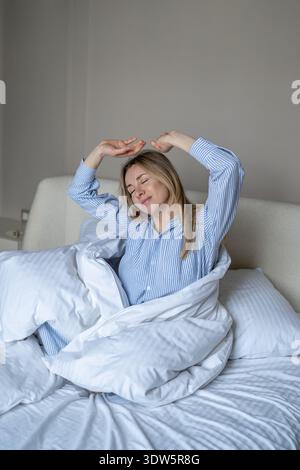 Una donna matura e felice che si sveglia a letto, si allunga con un sorriso e si gode di un'atmosfera tranquilla al mattino Foto Stock