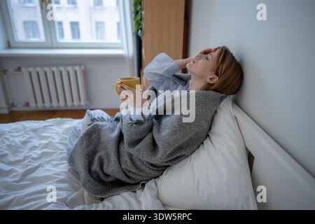 La donna malata esausta tocca la fronte calda a casa soffre di brividi, febbre nella stagione fredda, sintomi influenzali Foto Stock