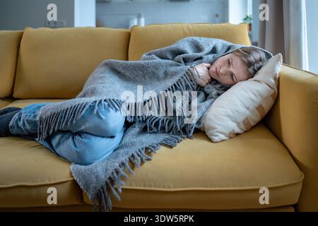 Donna malata con espressione dolorosa avvolta in plaid di lana sul divano con primi segni di raffreddore e influenza. Foto Stock