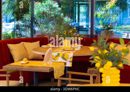 Un vivace ristorante interno presenta un tavolo rotondo in legno adornato con motivi al limone e vasi gialli, completato da lussuosi tavoli rossi a banchetto Foto Stock
