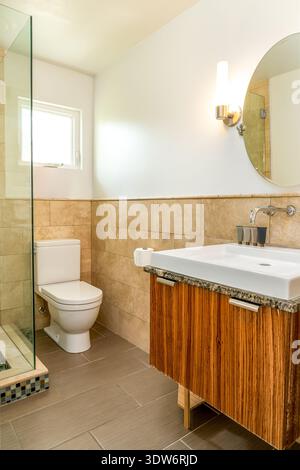 Bagno moderno con doccia in vetro, mobile galleggiante in legno, lavandino, specchio rotondo e WC Foto Stock
