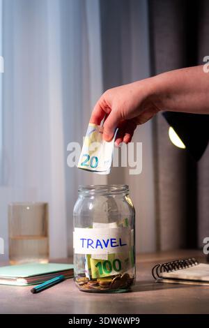 Versare il denaro contante in un vaso di vetro sul tavolo. Primo piano delle dita che inseriscono il denaro in un contenitore trasparente per viaggiare. Pianificazione finanziaria e. Foto Stock