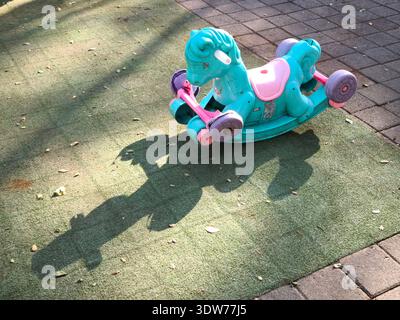 Un colorato giocattolo a dondolo per bambini si trova da solo su un parco giochi all'aperto, gettando una lunga ombra alla luce del sole del mattino Foto Stock