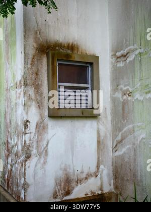 Una finestra chiusa e trascurata in un muro incrinato, usurato e ammuffito nell'angolo di un edificio Foto Stock