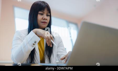Donna d'affari asiatica concentrata che usa la penna stilo con il computer portatile in ufficio. Responsabile femminile professionista che lavora su progetti creativi digitali o firma di documentari Foto Stock