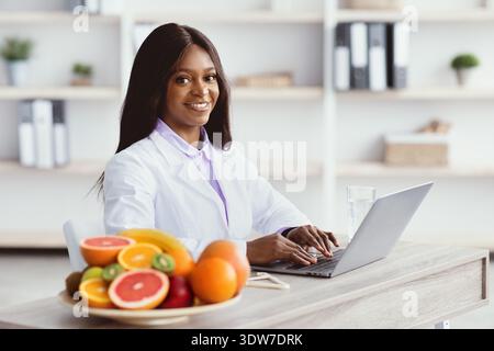 Nutrizionista che lavora su un laptop con frutta davanti presso Bright Office Space Foto Stock