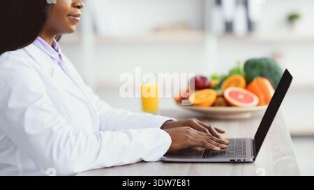 Nutrizionista che lavora su un computer portatile con frutta e succo sul tavolo in una stanza luminosa Foto Stock