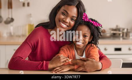 Madre e figlia condividono un momento di gioia mentre si siedono al tavolo della cucina durante il giorno Foto Stock