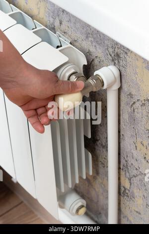 Una mano umana ruota la maniglia della valvola su un radiatore bianco liscio, regolando la temperatura nell'impianto di riscaldamento. Foto Stock