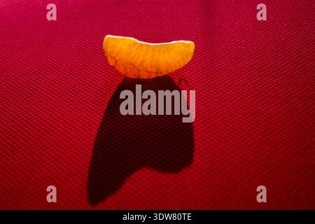 Una fotografia minimalista in stile pop-art di un singolo segmento di mandarino sbucciato adagiato su una superficie in tessuto rosso testurizzato, che crea una chiara ombra scura Foto Stock