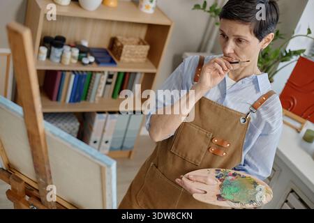 Donna caucasica di mezza età dipingere su tela mentre si pensa in classe d'arte Foto Stock