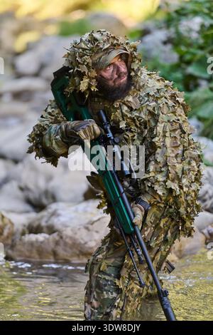 Cecchino soldato in Ghillie Suit con Rifle Crossing Stream durante l'operazione militare tattica mimetica Foto Stock
