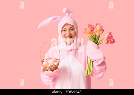 Divertente donna anziana in costume da coniglietto con cesto con uova di Pasqua e tulipani su sfondo rosa Foto Stock